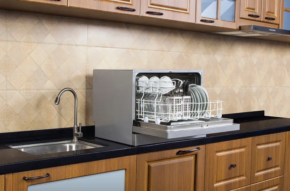A compact countertop dishwasher sitting on a small rental apartment counter next to a stainless sink, with a pull-out faucet visible and limited prep space