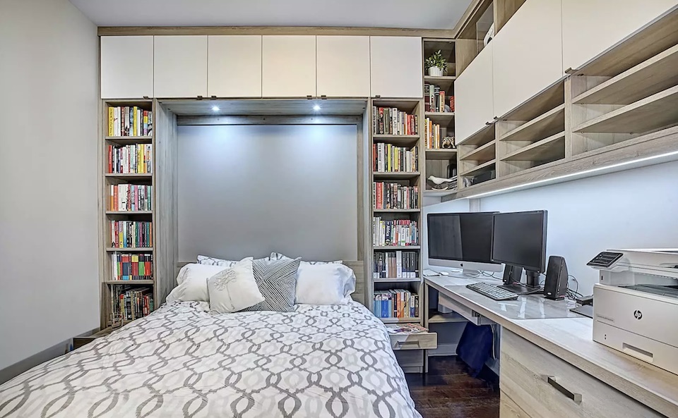 A small room with a Murphy bed closed against one wall and a tidy desk setup directly across from it, with a task chair and minimal wall art